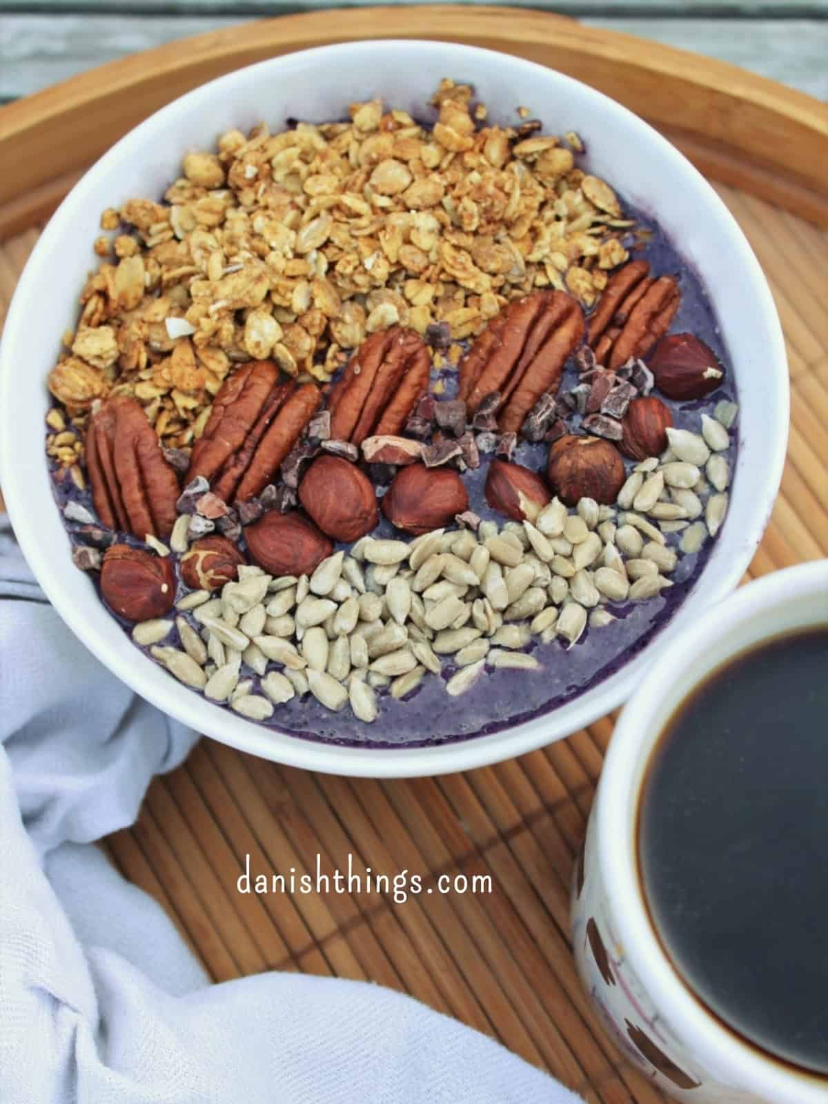 Sprød hverdagsgranola sødet med banan - granola uden tilsat sukker Blåbærsmoothiebowl toppet med granola, frø og nødder på en bakke med en kop kaffe. Lav en velsmagende blåbærsmoothie og top den med nødder og sprød granola sødet med banan. Få opskriften på både blåbærsmoothie og hverdagsgranola uden tilsat sukker på danishthings.com © Christel Parby #DanishThings #granola #smoothiebowl #blåbær #blåbærsmoothie #morgenmad #udentilsatsukker