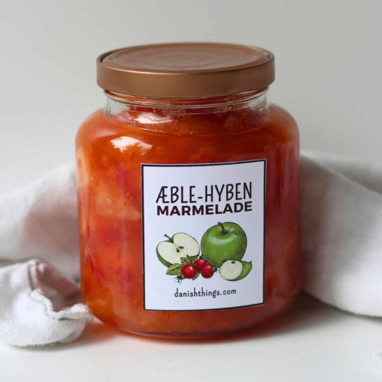 Hjemmelavet æblemarmelade med hyben i et glas med etiket på hvid baggrund. Æblemarmelade med hyben. Få opskriften på den lækreste æblemarmelade med hyben! En skøn efterårsmarmelade, hvor hyben giver smag og farve, og æblerne giver fylde. Lav marmeladen og print de gratis etiketter – find det hele på danishthings.com © Christel Parby #DanishThings #æblemarmelade #hybenmarmelade #hjemmelavet #efterårsmad #sanke