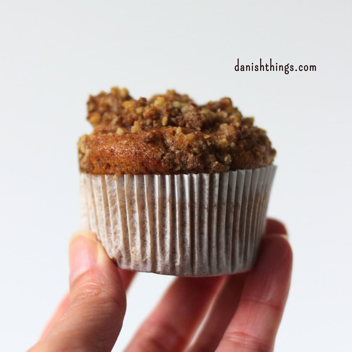 En friskbagt bananmuffin med sprød valnøddecrumble i en hvid papirform, holdt op foran en lys baggrund. Bananmuffinsene kan bages med crumble eller pyntes med glasur og creme. Find opskriften på Danish Things © Christel Parby #DanishThings #bananmuffins #banan #muffins #bananopskrift #muffinmetoden #hjemmebag #valnøddecrumble #valnødder #brunebananer #modnebananer #kanel #kagekrydderi #kaffeglasur #chantillycreme #muffintopping #crumble #stopmadspild