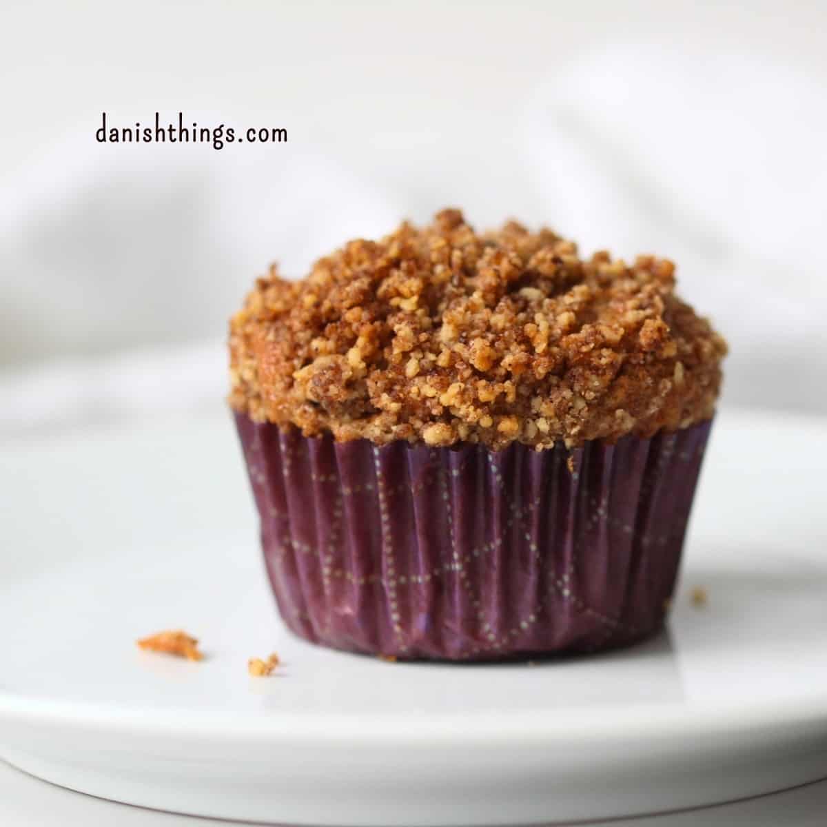 Færdigbagt bananmuffin med gylden valnøddecrumble i en lilla papirform på en hvid tallerken. Crumblen med kagekrydderi giver ekstra sødme og en sprød topping til den bløde muffin. Find opskriften på Danish Things © Christel Parby #DanishThings #bananmuffins #banan #muffins #bananopskrift #muffinmetoden #hjemmebag #valnøddecrumble #valnødder #modnebananer 