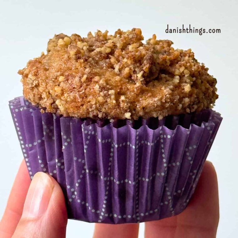 Saftig bananmuffin med valnøddecrumble i en lilla papirform, holdt op foran en lys baggrund. Muffinen er bagt med modne bananer, kanel og grofthakkede valnødder. Find opskriften på Danish Things © Christel Parby #DanishThings #bananmuffins #banan #muffins #bananopskrift #muffinmetoden #hjemmebag #valnøddecrumble #valnødder #modnebananer #kanel #kagekrydderi #crumble #stopmadspild