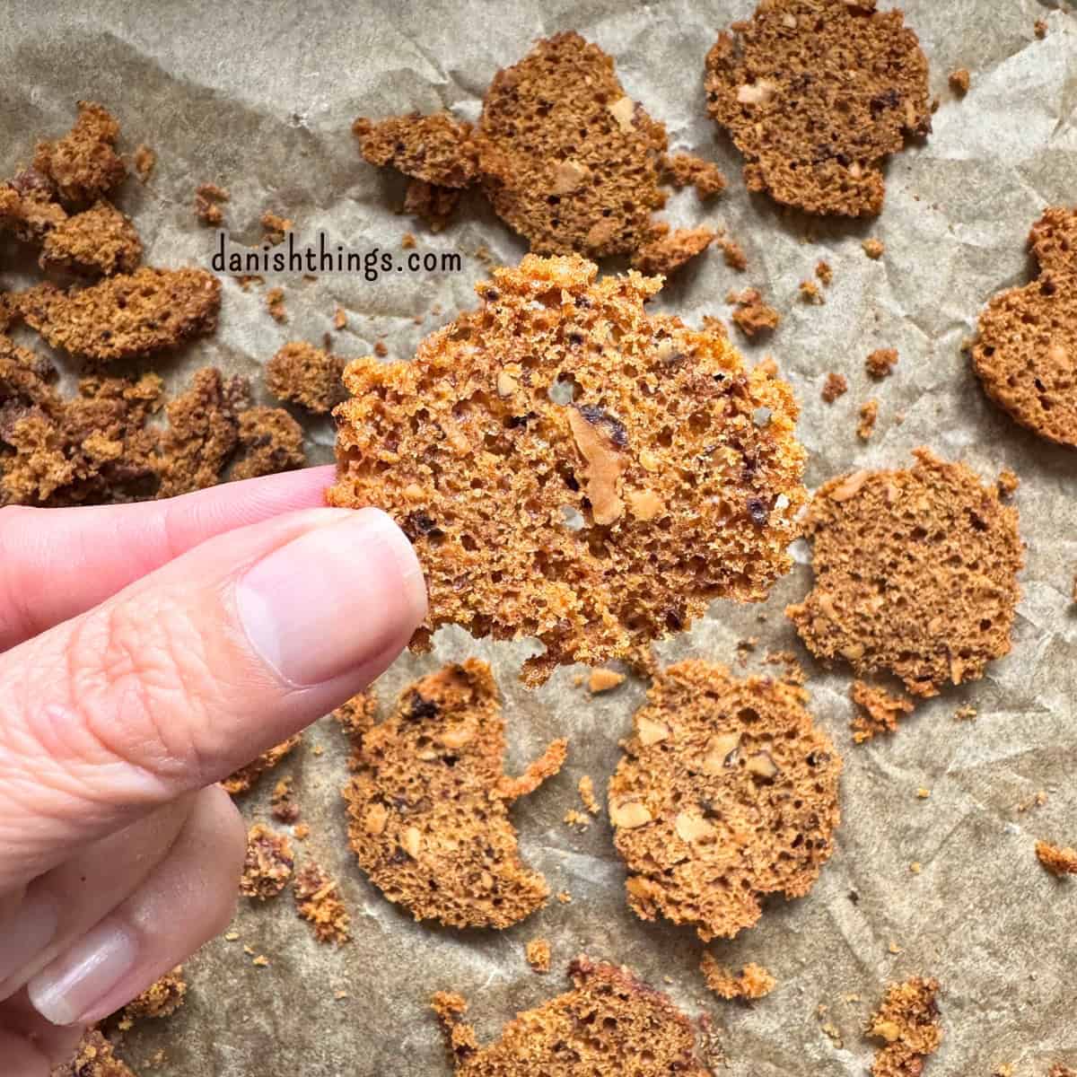 Tørrede skiver af bananmuffins med valnødder på bagepapir, hvor en skive holdes op for at vise den sprøde struktur. Muffins tilovers kan skæres i skiver og tørres i ovnen til en sprød snack eller kagerasp. Find opskriften på Danish Things © Christel Parby #DanishThings #bananmuffins #banan #muffins #bananopskrift #brunebananer #modnebananer #stopmadspild