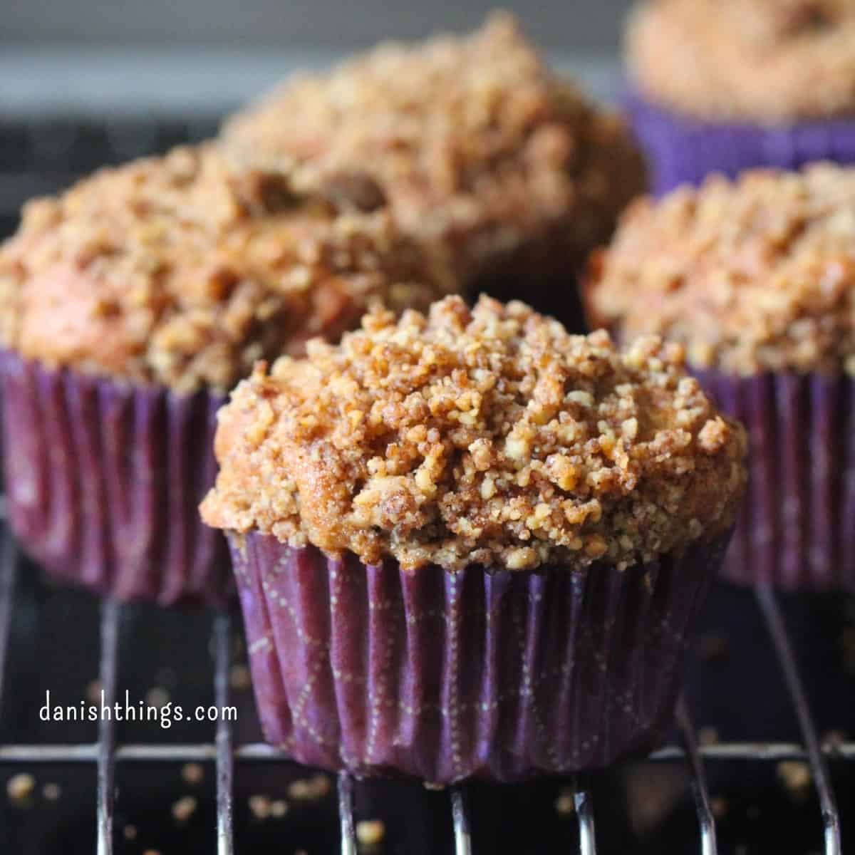 Friskbagte bananmuffins med valnødder og sprød crumble i lilla papirforme på en bagerist. De saftige muffins er lavet med modne bananer og toppet med en sød valnøddecrumble med kagekrydderi. Find opskriften på Danish Things © Christel Parby #DanishThings #bananmuffins #banan #muffins #bananopskrift #muffinmetoden #hjemmebag #valnøddecrumble #valnødder #brunebananer #kanel #kagekrydderi #crumble #stopmadspild