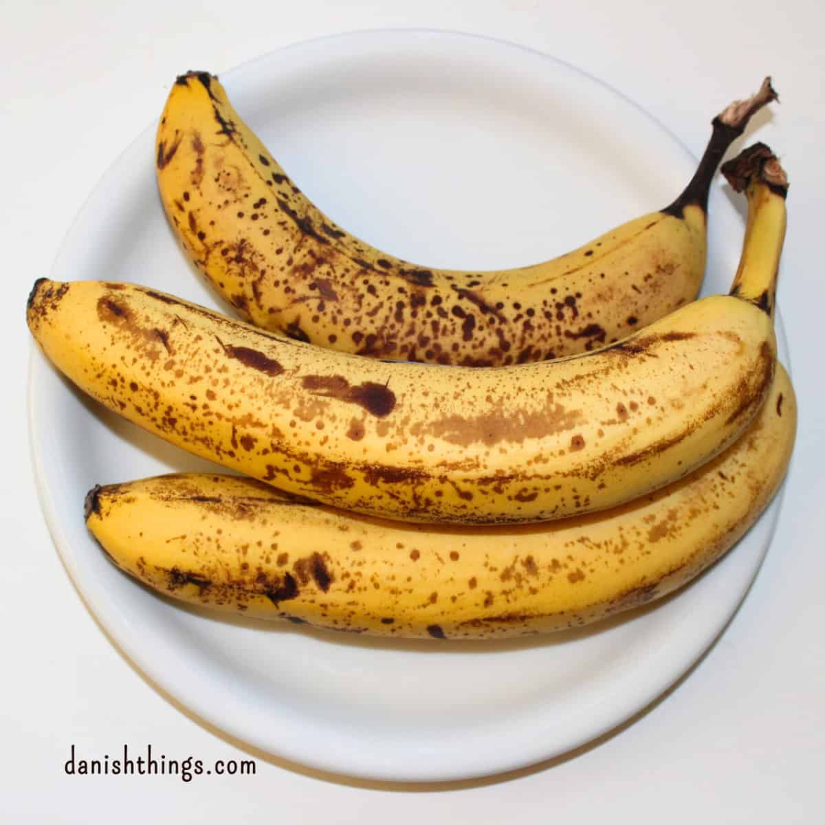 3 modne bananer med masser af brune pletter liggende på en hvid tallerken. Brug dine brune bananer til at bage bløde og saftige muffins, til granola og banankager – stop-madspild. Find opskriften på Danish Things © Christel Parby #DanishThings #bananmuffins #banan #bananopskrift #muffinmetoden #banankage #hjemmebag #modnebananer #brunebananer #stopmadspild