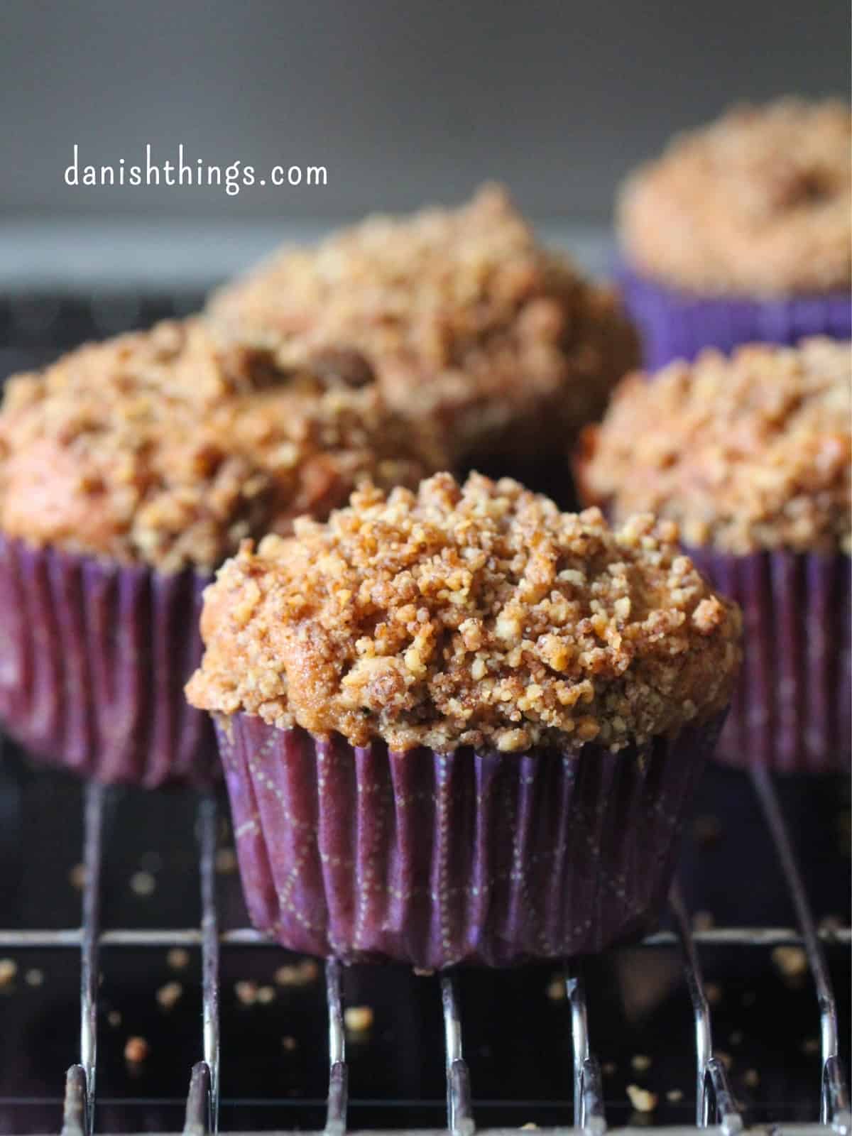 Friskbagte bananmuffins med valnødder og sprød crumble i lilla papirforme på en bagerist. De saftige muffins er lavet med modne bananer og toppet med en sød valnøddecrumble med kagekrydderi. Find opskriften på Danish Things © Christel Parby #DanishThings #bananmuffins #banan #muffins #bananopskrift #muffinmetoden #hjemmebag #valnøddecrumble #valnødder #brunebananer #kanel #kagekrydderi #crumble #stopmadspild