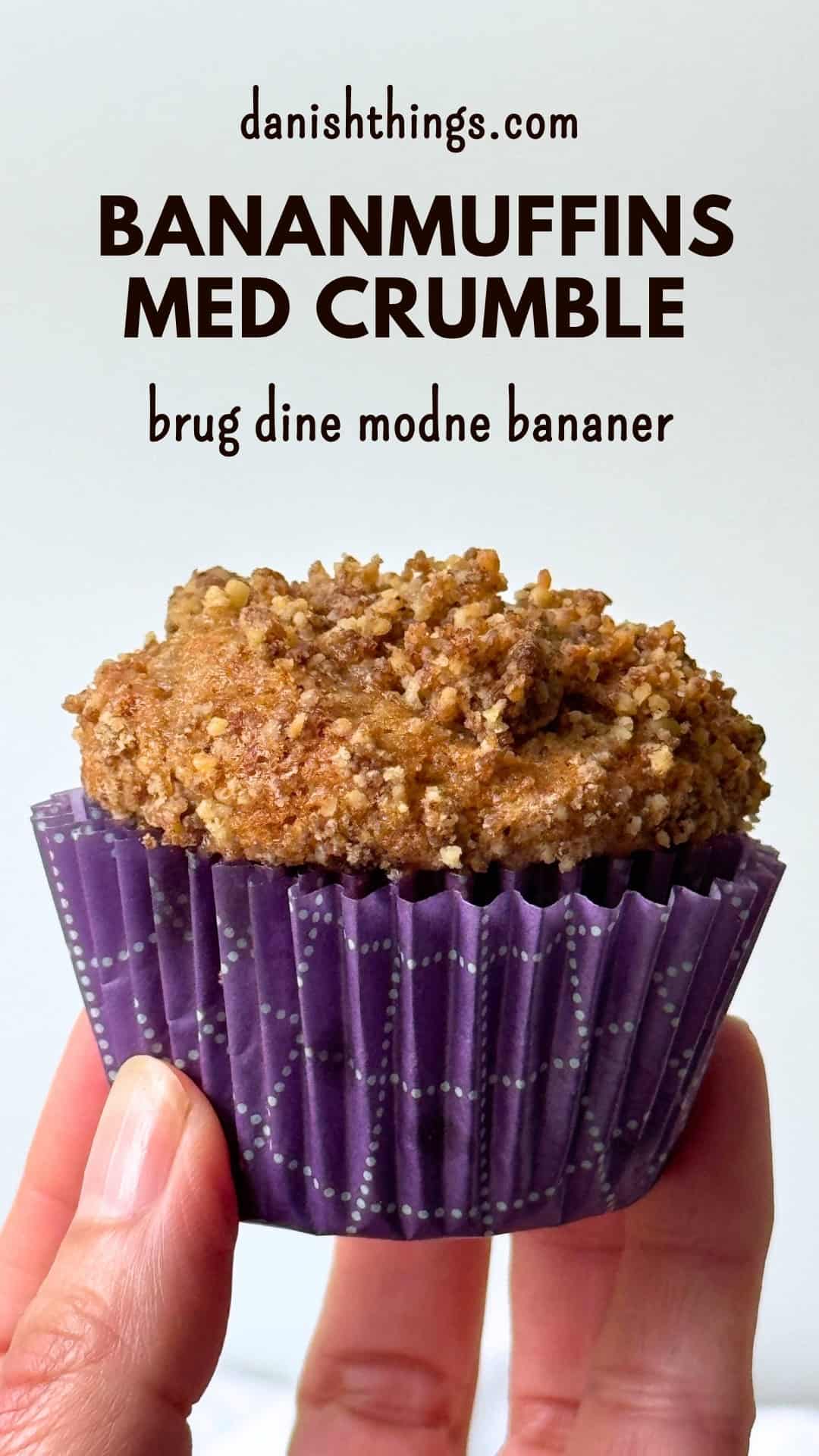 Bananmuffin med gylden valnøddecrumble i en lilla papirform, holdt op foran en lys baggrund. Bananmuffins med crumble – brug dine modne bananer til bløde og saftige muffins med valnødder og en sprød topping med kagekrydderi. Find opskriften på Danish Things © Christel Parby #DanishThings #bananmuffins #banan #muffins #bananopskrift #muffinmetoden #hjemmebag #valnøddecrumble #valnødder #brunebananer #kanel #kagekrydderi #crumble #stopmadspild