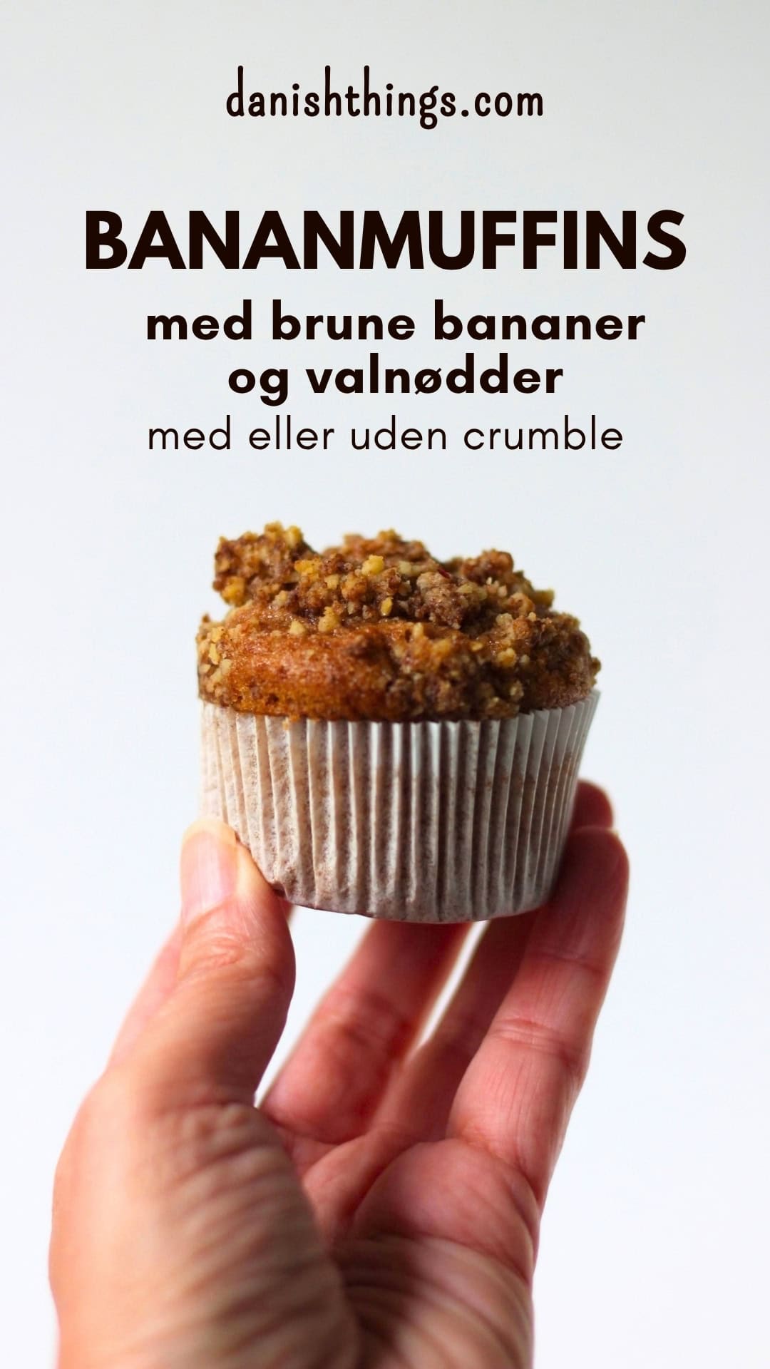 Bananmuffin med valnøddecrumble i en hvid papirform, holdt op foran en lys baggrund. Bananmuffins med brune bananer og valnødder – bløde og saftige muffins, som er nemme at bage med eller uden crumble. Find opskriften på Danish Things © Christel Parby #DanishThings #bananmuffins #banan #muffins #bananopskrift #muffinmetoden #hjemmebag #valnøddecrumble #valnødder #brunebananer #kanel #kagekrydderi #muffintopping #crumble #stopmadspild