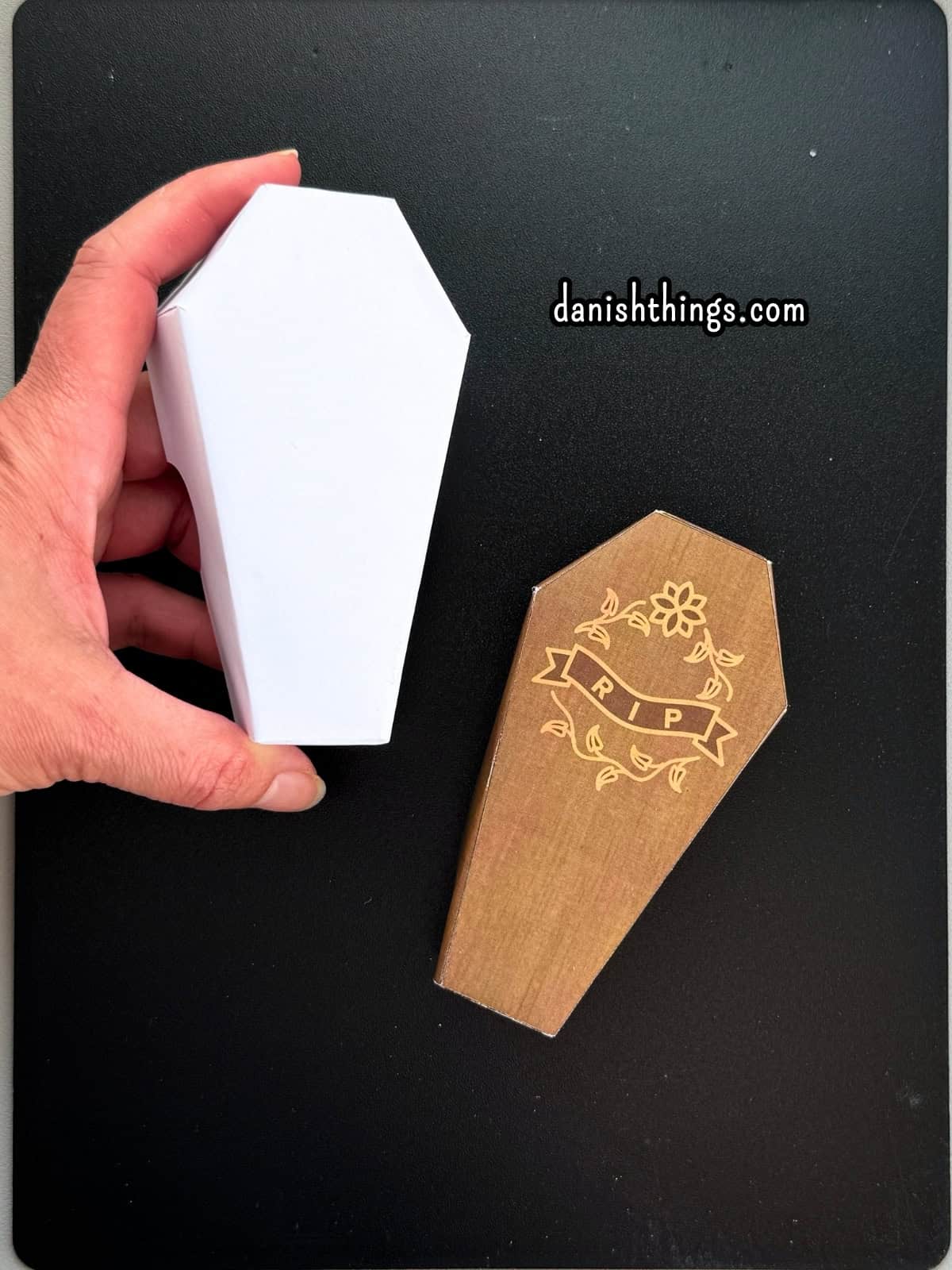 En hånd holder en hvid papirkiste ved siden af en brun kiste med RIP-motiv på låget. Billedet viser forskellen mellem den hvide udekorerede halloweenkiste og den farvede udgave af kisten.
© Christel Parby · danishthings.com · #DanishThings #Halloweenkiste #PrintSelv #Papirpynt
