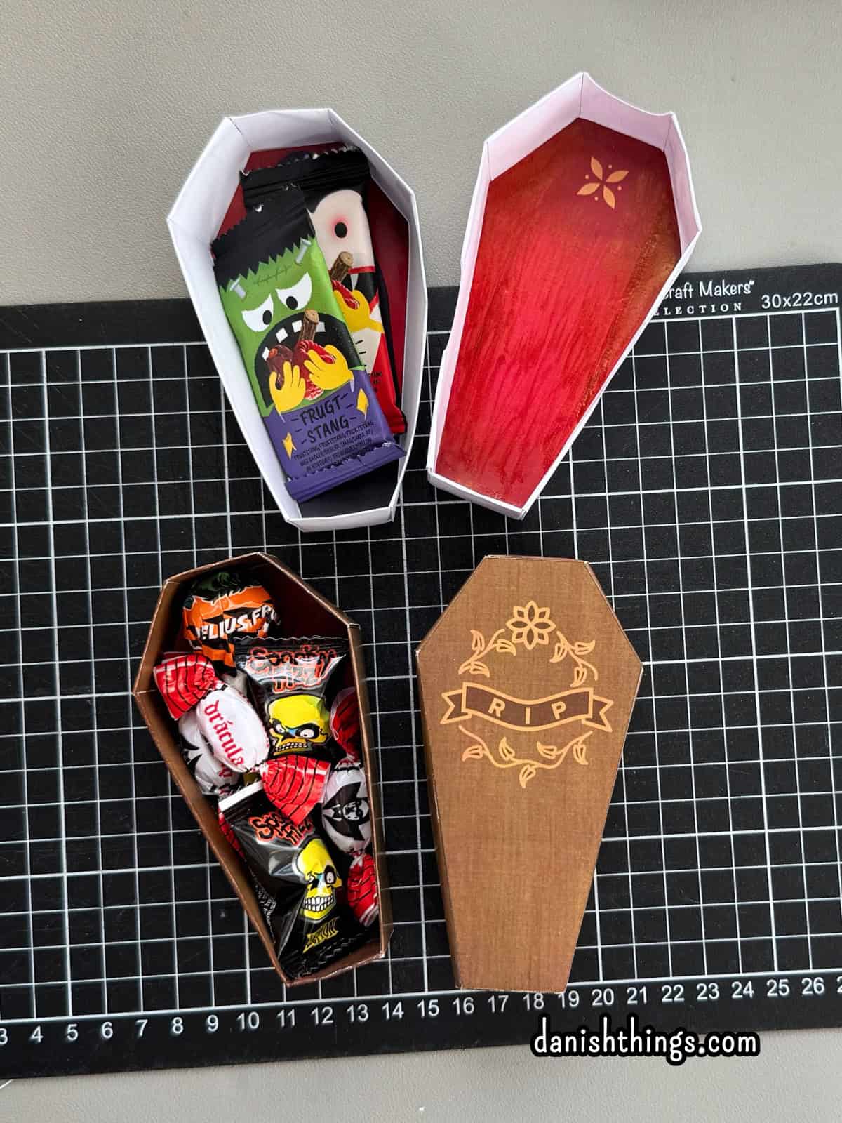 To kister af papir viser forskellige muligheder for dekoration og fyld til halloween, fx slik og frugtstænger. De brune og hvide kister kan bruges som pynt eller gaveæsker.
© Christel Parby · danishthings.com · #DanishThings #Halloweenpynt #Gaveæske #Papirkiste