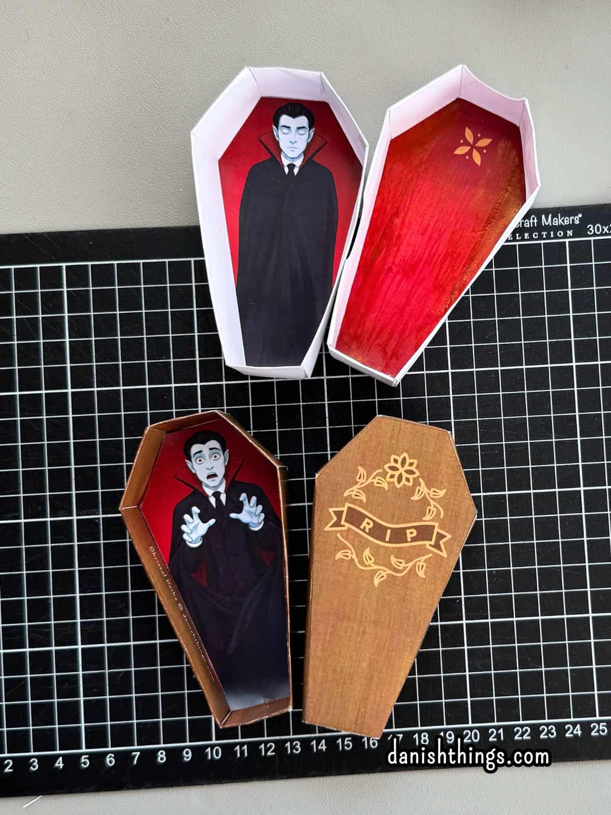 To papirkister ligger på et sort skæreunderlag. Den hvide kiste har røde indersider og en sovende vampyr, mens den brune kiste har en forskrækket vampyr og et RIP-låg.
© Christel Parby · danishthings.com · #DanishThings #Halloweenkiste #Vampyr #GratisPrint