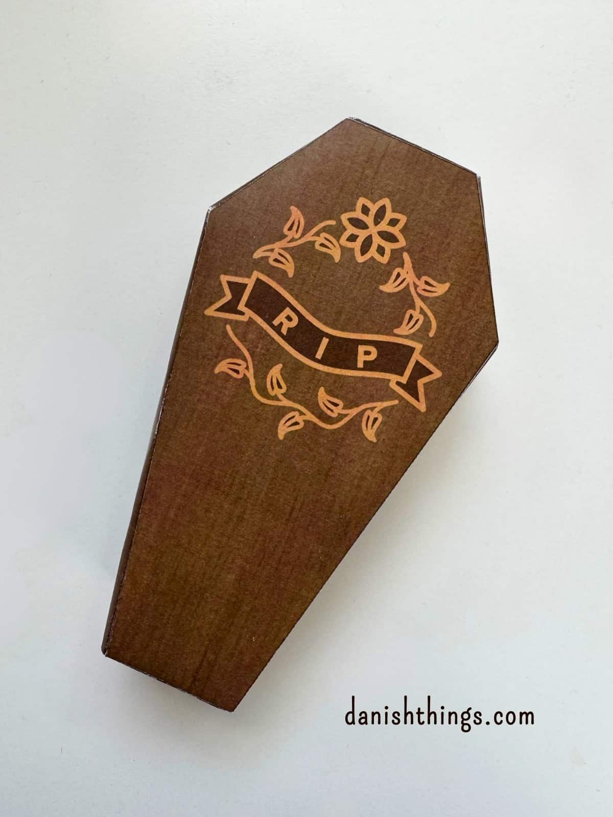 En brun kiste, hvor låget er dekoreret med et RIP-motiv, ligger på en lys baggrund. Kisten er samlet og klar til at anvende som dekoration eller gaveæske.
© Christel Parby · danishthings.com · #DanishThings #Halloweenpynt #Kistelåg #Papirpynt