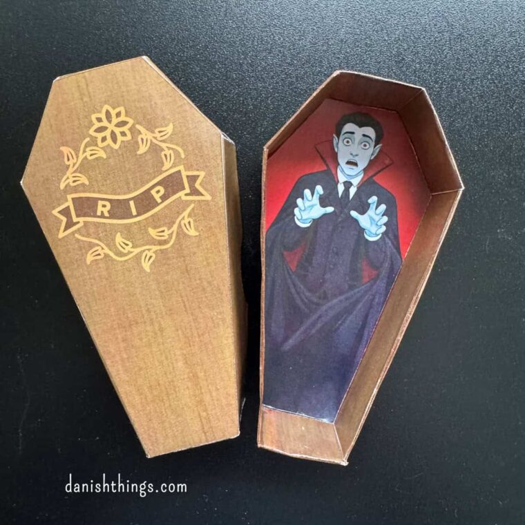 Brun halloween kiste af papir med et låg dekoreret med RIP og blomster. I den åbne kiste ligger en forskrækket vampyr med hænderne løftet. Lav din egen kiste med gratis print fra Danish Things. © Christel Parby · danishthings.com · #DanishThings #Halloweenkiste #Halloweenpynt #GratisPrint