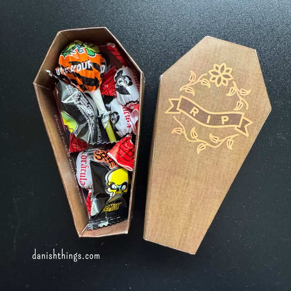 En brun kiste af papir er fyldt med indpakket halloweenslik. Det matchende låg med RIP-motiv ligger ved siden af. Kisten kan bruges som en lille gaveæske eller som dekoration til Halloween.
© Christel Parby · danishthings.com · #DanishThings #HalloweenGave #Gaveæske #Halloweenpynt