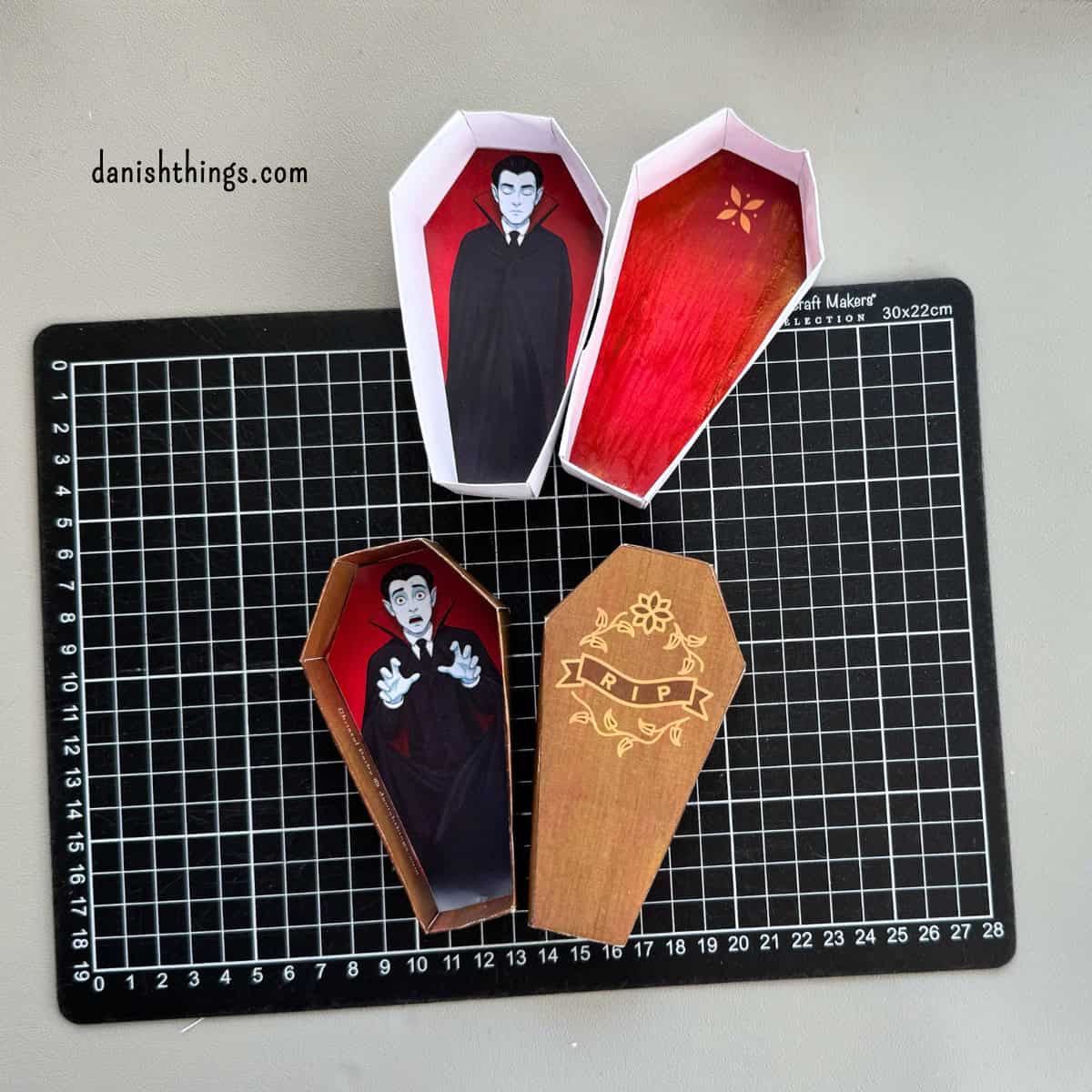 To papirkister ligger på et sort skæreunderlag. Den hvide kiste har røde indersider og en sovende vampyr, mens den brune kiste har en forskrækket vampyr og et RIP-låg.
© Christel Parby · danishthings.com · #DanishThings #Halloweenkiste #Vampyr #GratisPrint
