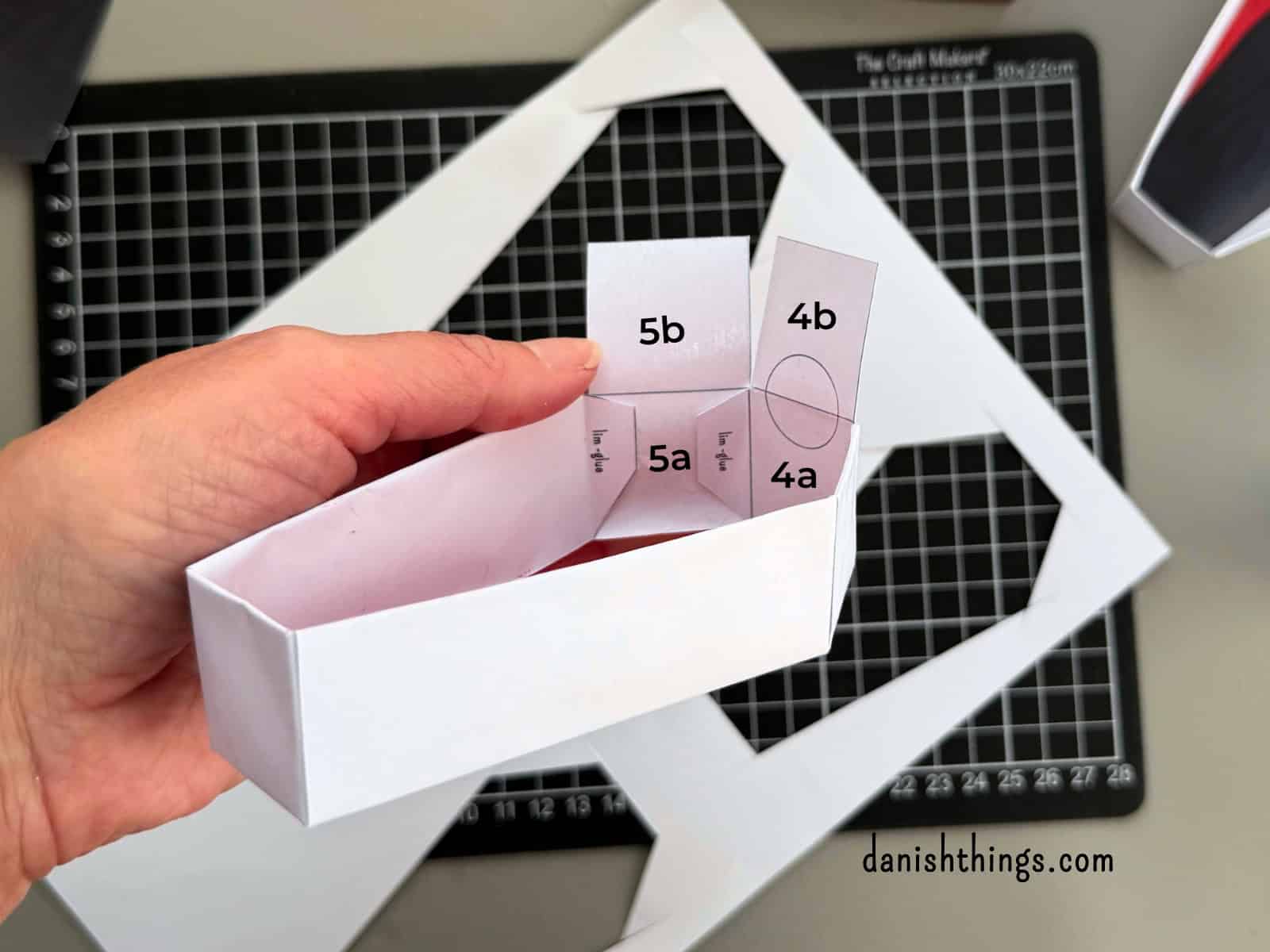 En hånd holder et næsten færdigsamlet hvidt kistelåg. De nummererede dele og limflapper ved hovedenden er synlige, så man kan følge samlingen af de sidste sider. © Christel Parby · danishthings.com · #DanishThings #Kistelåg #Samlevejledning #HalloweenDIY