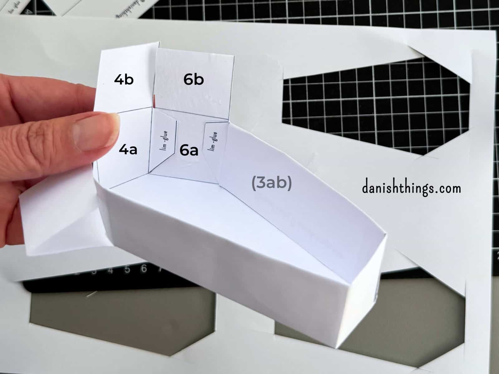 En hånd holder en hvid kistebund, hvor delene 4a/4b og 6a/6b er synlige, og 3a/3b er sat i parentes. Billedet hjælper med at finde de rigtige dele under samlingen af de sidste vægge.
© Christel Parby · danishthings.com · #DanishThings #Kistebund #Samlevejledning #GratisPrint