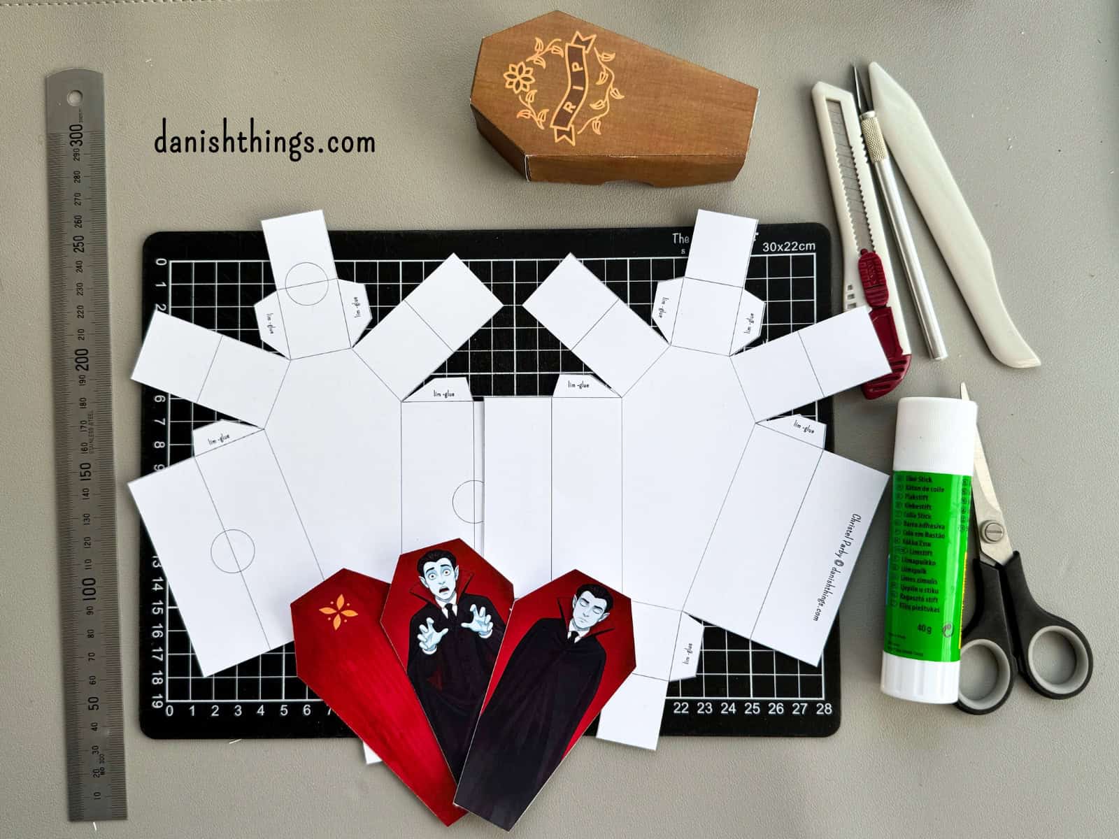 Udklippede hvide kistedele og forskellige printede vampyrer ligger på et sort skæreunderlag sammen med redskaber til samlingen. Forberedelse af en halloweenkiste med gratis print fra Danish Things.
© Christel Parby · danishthings.com · #DanishThings #HalloweenDIY #KlipOgFold #GratisPrint
