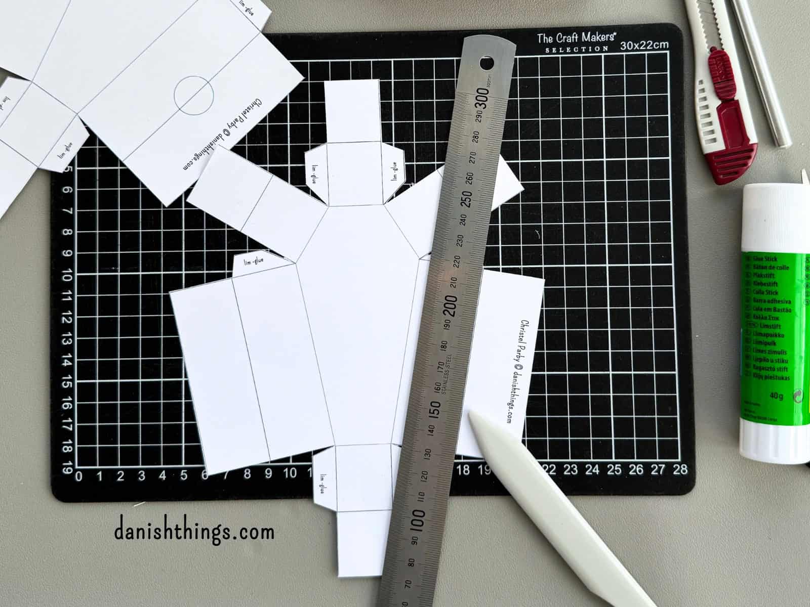 En hvid kiste ligger på et sort skæreunderlag med en metallineal og et falseben. Foldelinjer forberedes med falsebenet, inden papirkisten foldes og samles.
© Christel Parby · danishthings.com · #DanishThings #Papirkiste  #HalloweenDIY #Papirklip #GratisPrint
