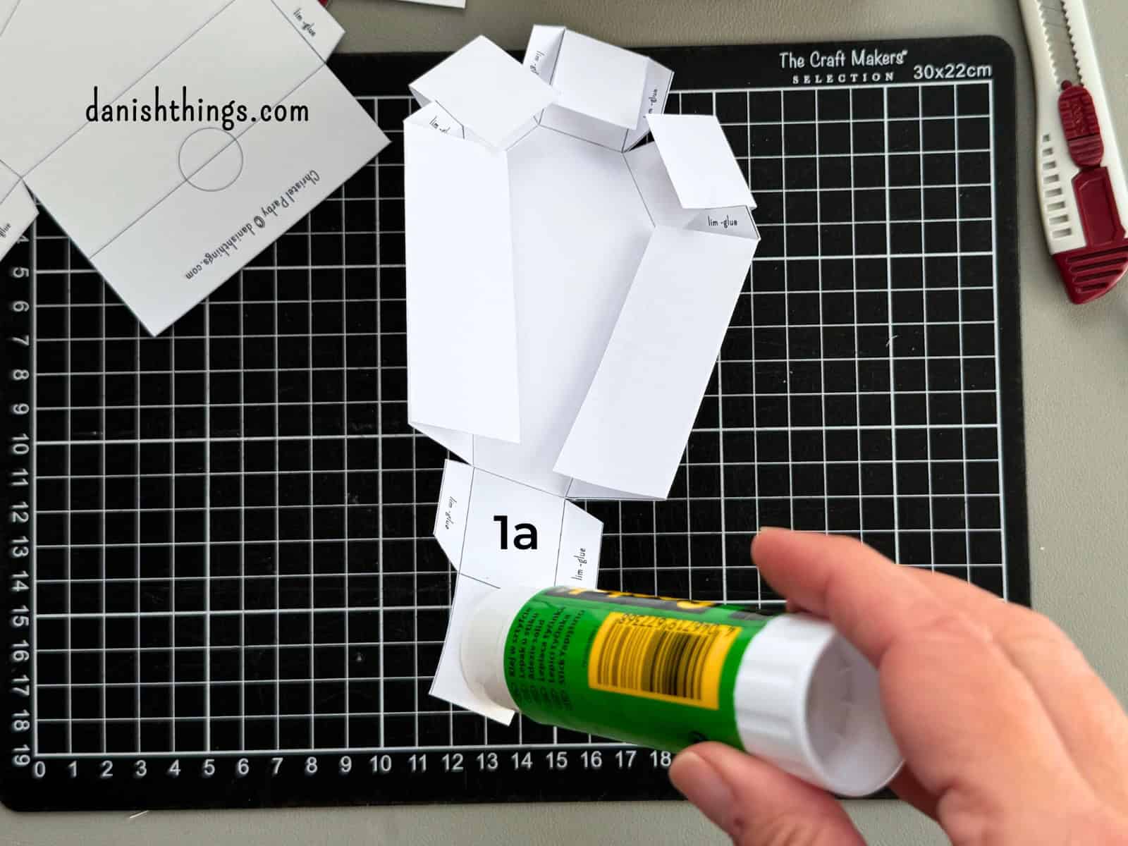 En limstift påfører lim på den hvide kistes fodende, hvor delene 1a og 1b skal samles. Limflapperne holdes fri, så de senere kan placeres mellem sidevæggene.
© Christel Parby · danishthings.com · #DanishThings #Kistebund #LimOgFold #HalloweenDIY