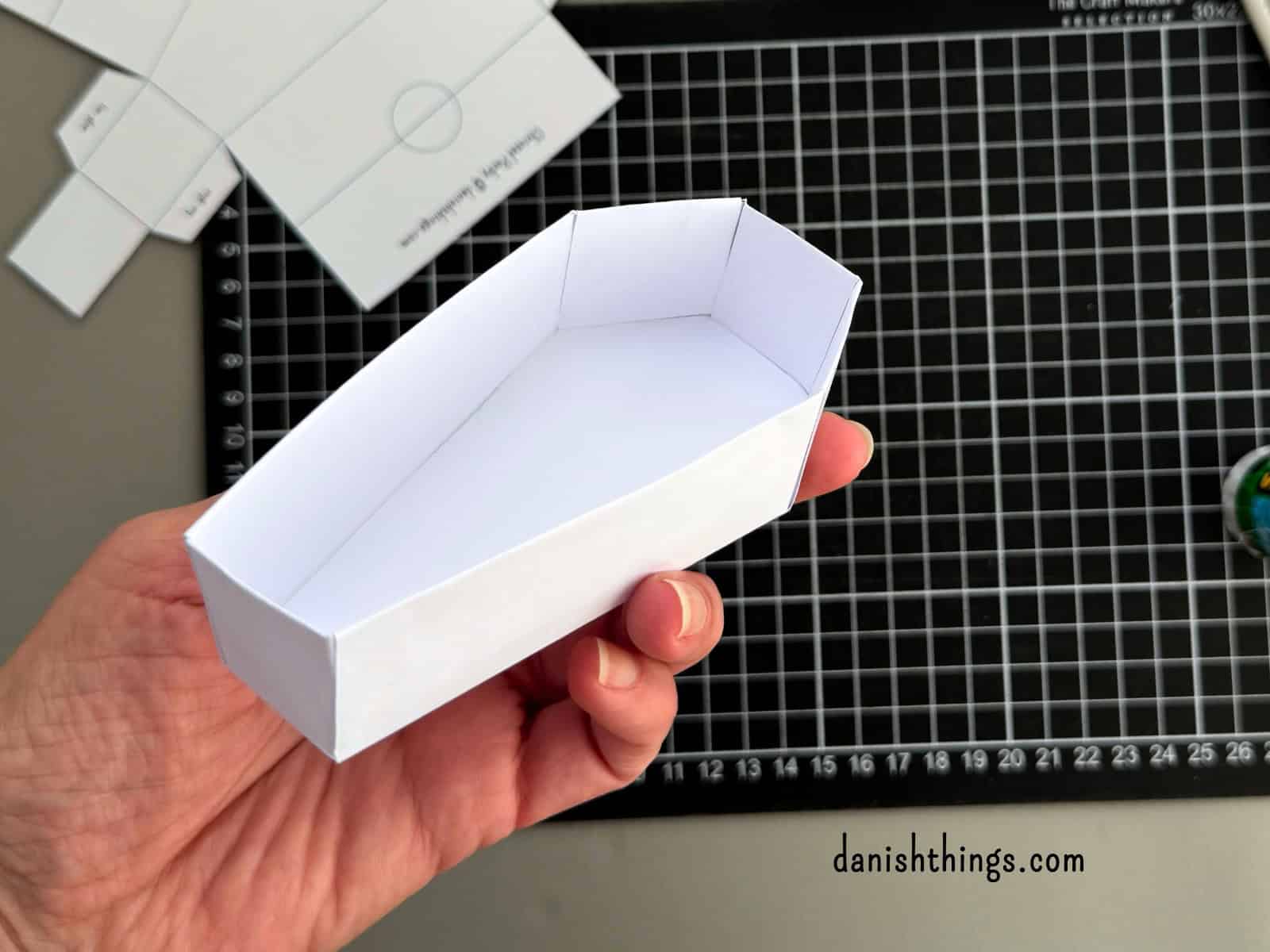 En samlet hvid kistebund holdes i hånden over et sort skæreunderlag. De høje, dobbelte sidevægge og den færdige kisteform kan ses.
© Christel Parby · danishthings.com · #DanishThings #Kistebund #HalloweenDIY #Papirpynt