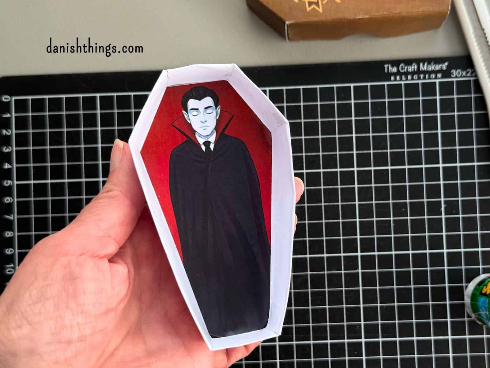 En hånd holder en færdigsamlet hvid kistebund med en sovende vampyr på rødt for. Kisten kan dekoreres indvendigt med et af de gratis print fra Danish Things. © Christel Parby · danishthings.com · #DanishThings #Halloweenkiste #Vampyr #GratisPrint
