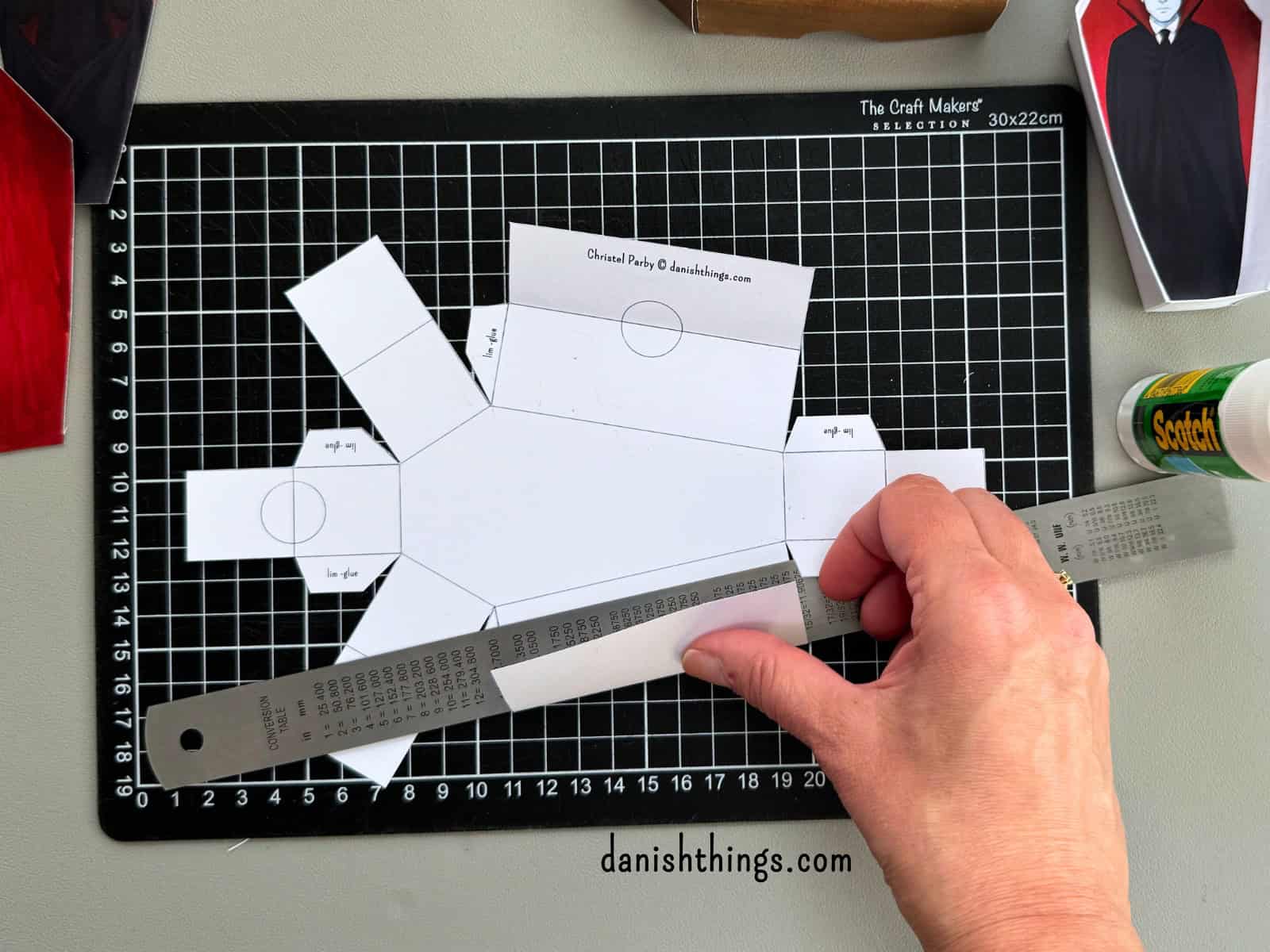 En hvid udklippet kiste ligger på et sort skæreunderlag med en metallineal. Linealen bruges til at folde papiret præcist langs de rette linjer, før kisten samles. 
© Christel Parby · danishthings.com · #DanishThings #Papirfoldning #HalloweenDIY #Kisteprint

